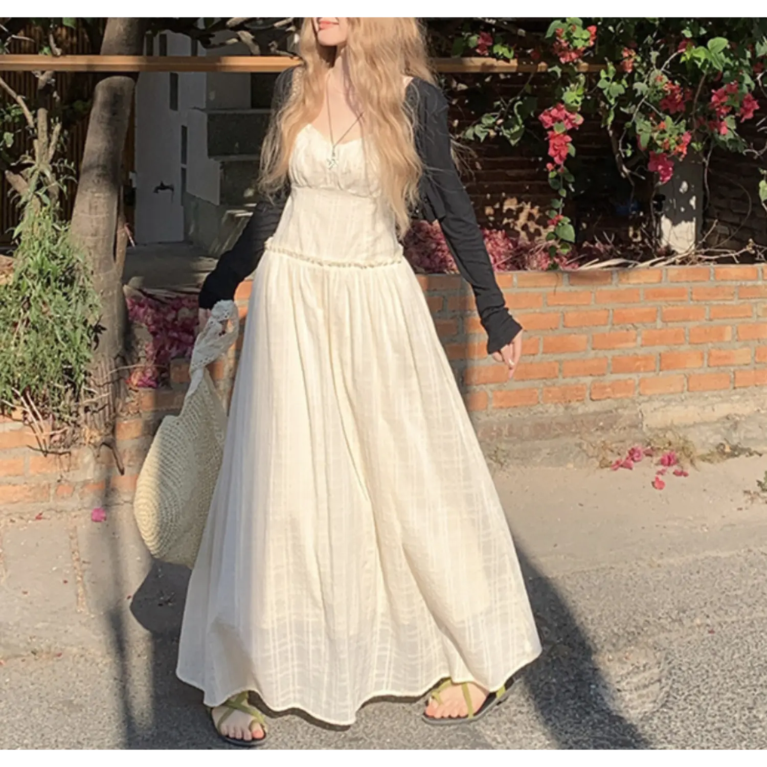 Outdoor brick wall full styling shot of cream textured slip maxi dress layered with slim long sleeve black knit cropped cardigan, ruched sweetheart neck, ruffle empire waist, sweeping flowy skirt, paired with woven straw shoulder bag, green slide sandals, blooming bougainvillea background