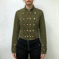 Model wearing olive green military cropped jacket, stand collar, rows of knot‑loop brass button decor, fitted silhouette, paired with black pants