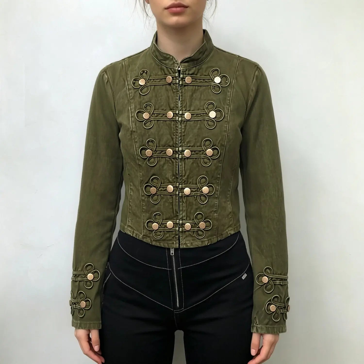 Model wearing olive green military cropped jacket, stand collar, rows of knot‑loop brass button decor, fitted silhouette, paired with black pants