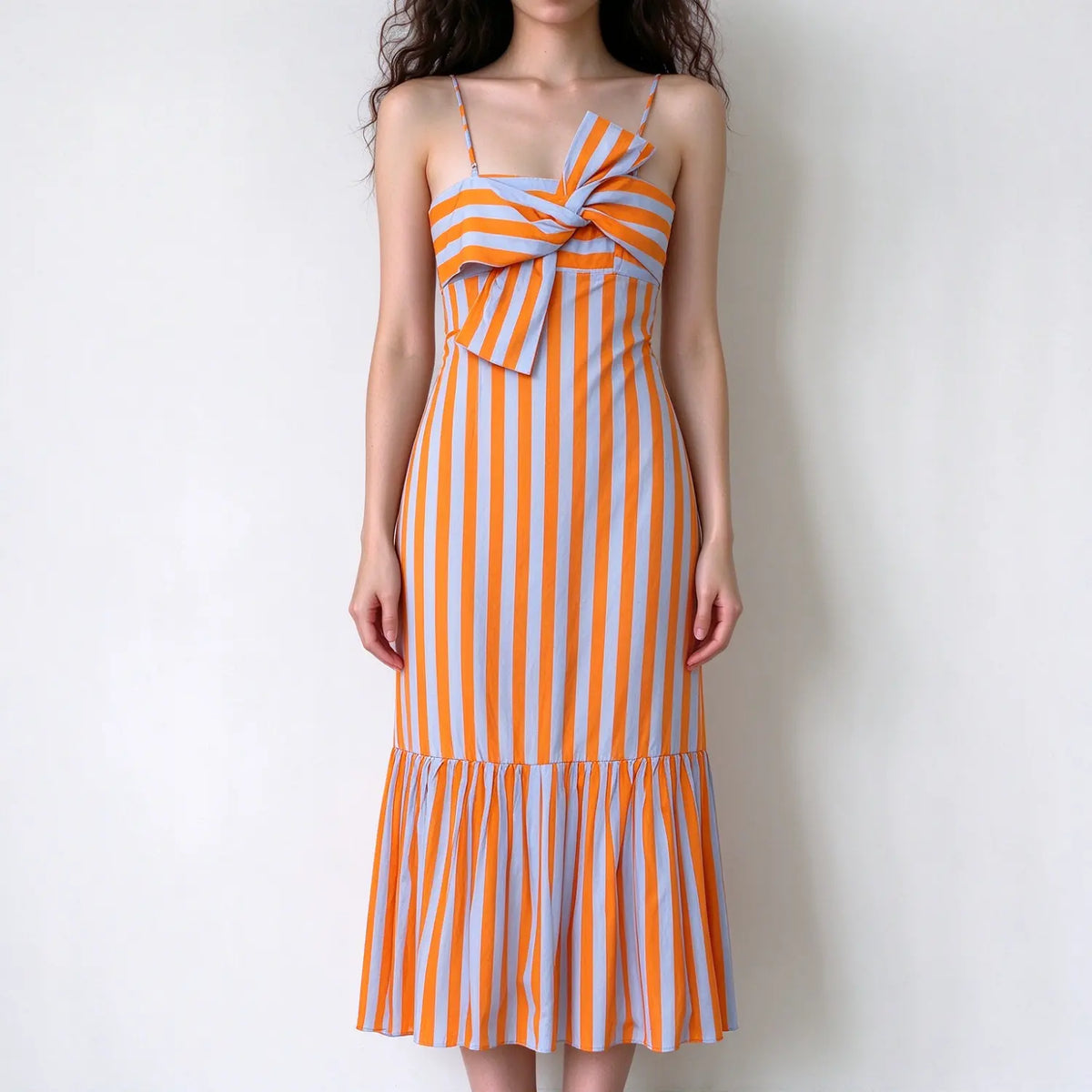 Model wearing orange and light blue vertical stripe maxi dress, thin spaghetti straps, large front bow decoration, ruffled maxi hem, solid white background