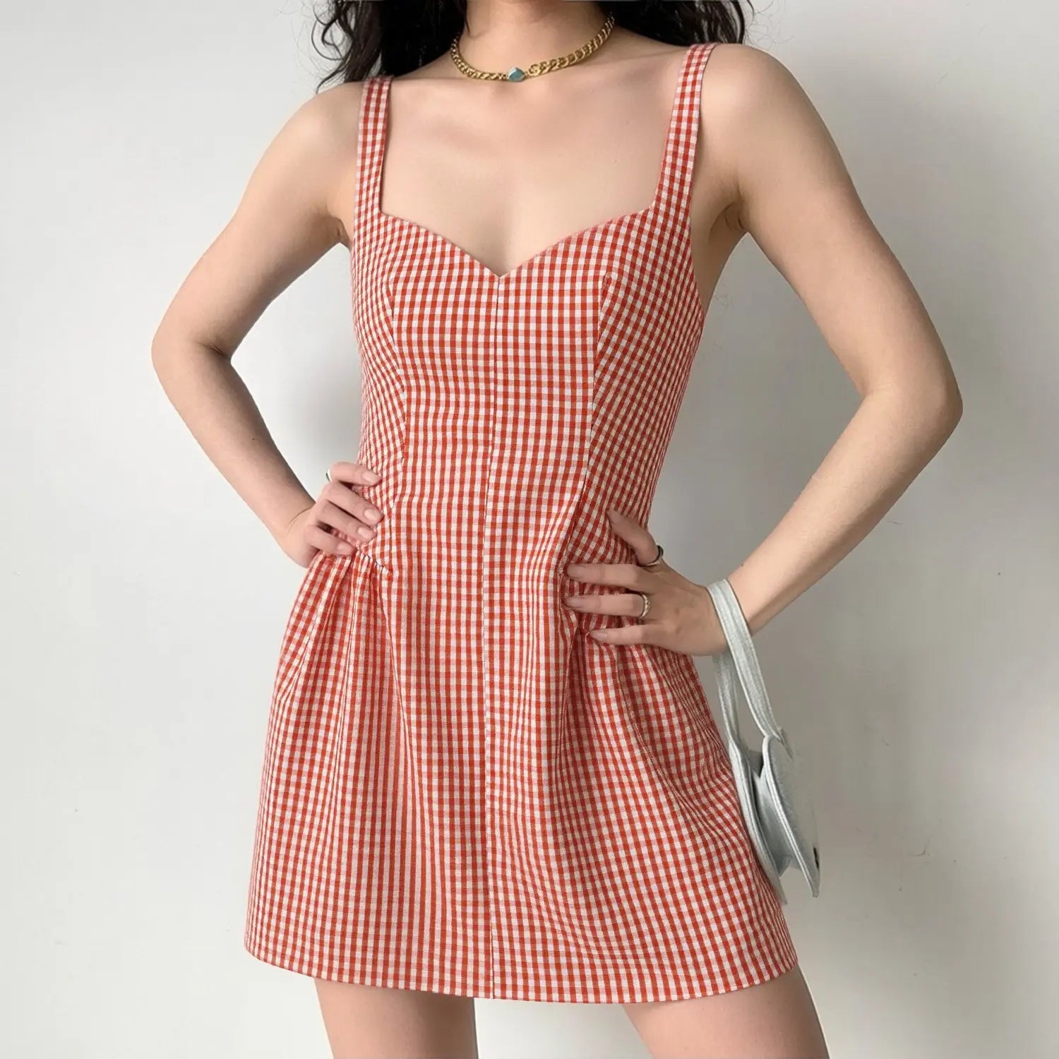 Lifestyle model shot wearing orange gingham sweetheart cami mini dress, paired with thin gold chain necklace and light gray mini handbag