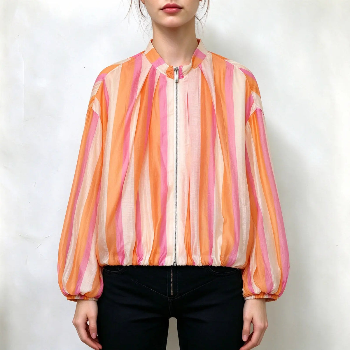 Model wearing sheer striped blouse, vertical orange pink cream stripes, stand collar, full front zipper, balloon long sleeves with elastic cuffs, loose billowy fit, paired with black jeans, plain white background