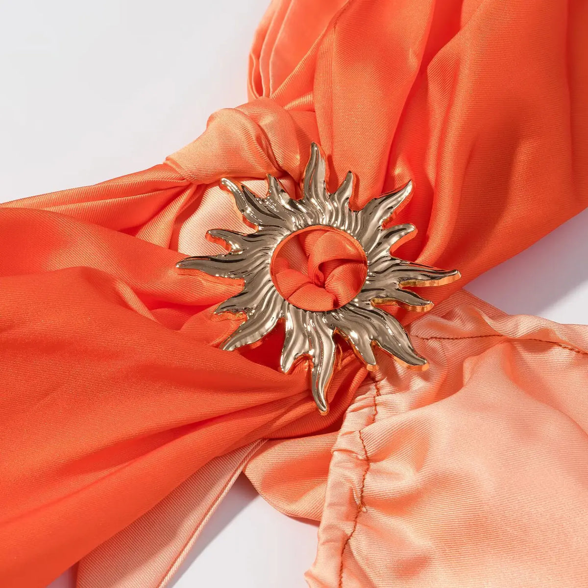 Macro shot of golden sun-shaped metal ornament on orange satin maxi dress, fabric knotted through hollow sun decor