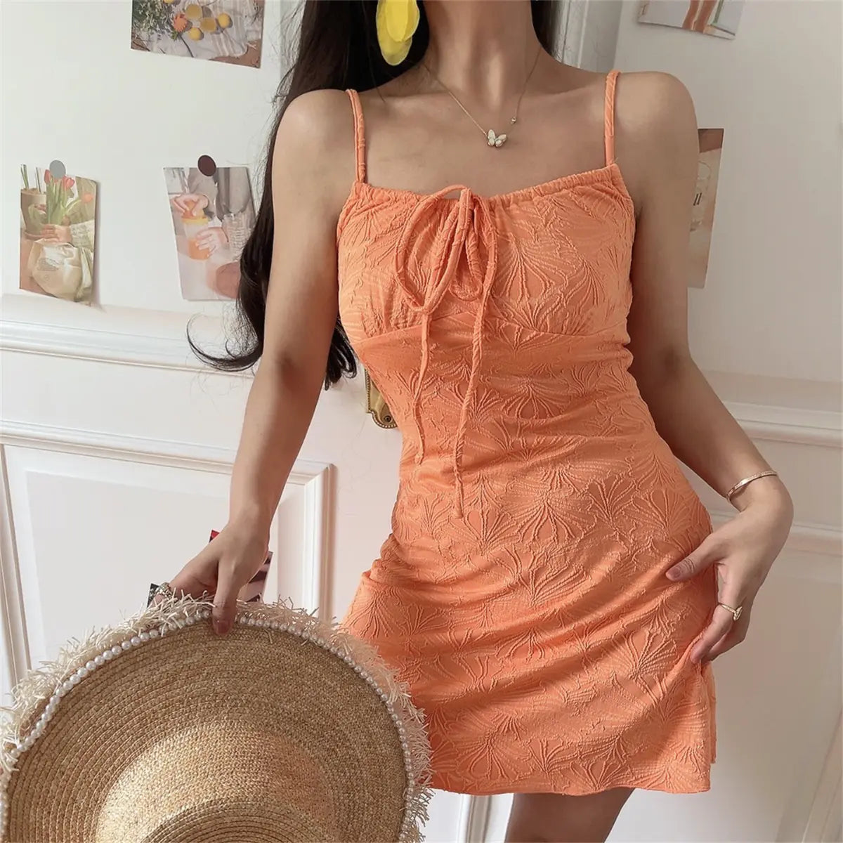 Woman wearing an orange textured mini dress with spaghetti straps, front tie detail, and embossed leaf pattern, holding a straw hat.
