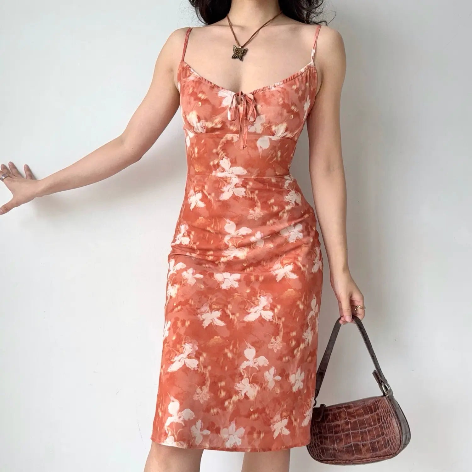 Lifestyle shot of woman wearing orange watercolor floral slip midi dress, thin spaghetti straps and front bust tie detail, paired with brown crocodile handbag and butterfly necklace