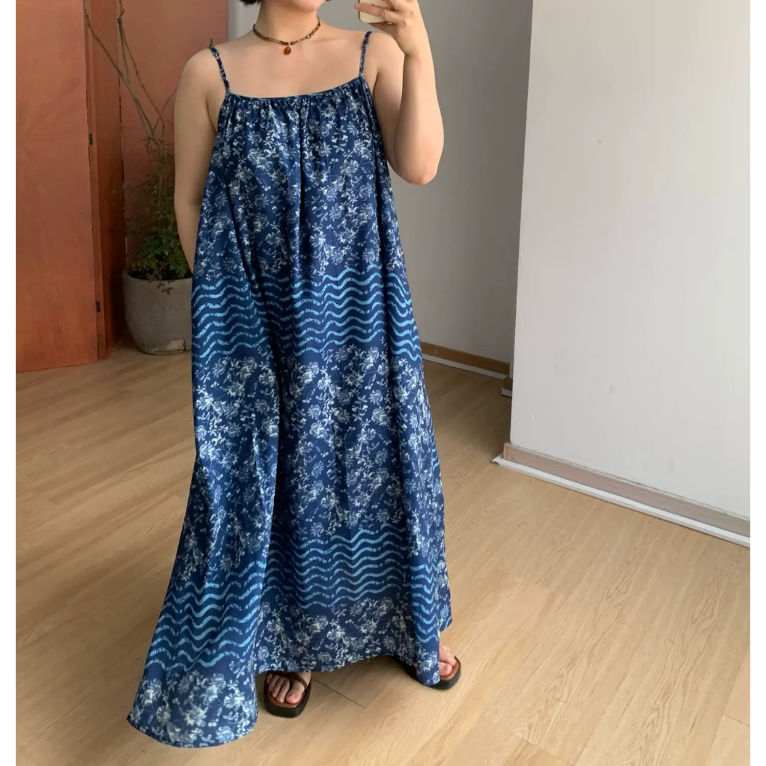Front indoor mirror full shot of loose fit batik cami maxi dress, gathered square neckline, thin matching spaghetti straps, segmented white floral & pale blue wave horizontal print panels, deep indigo base, hidden side pocket detail, brown platform slide sandals, minimalist light wood home interior
