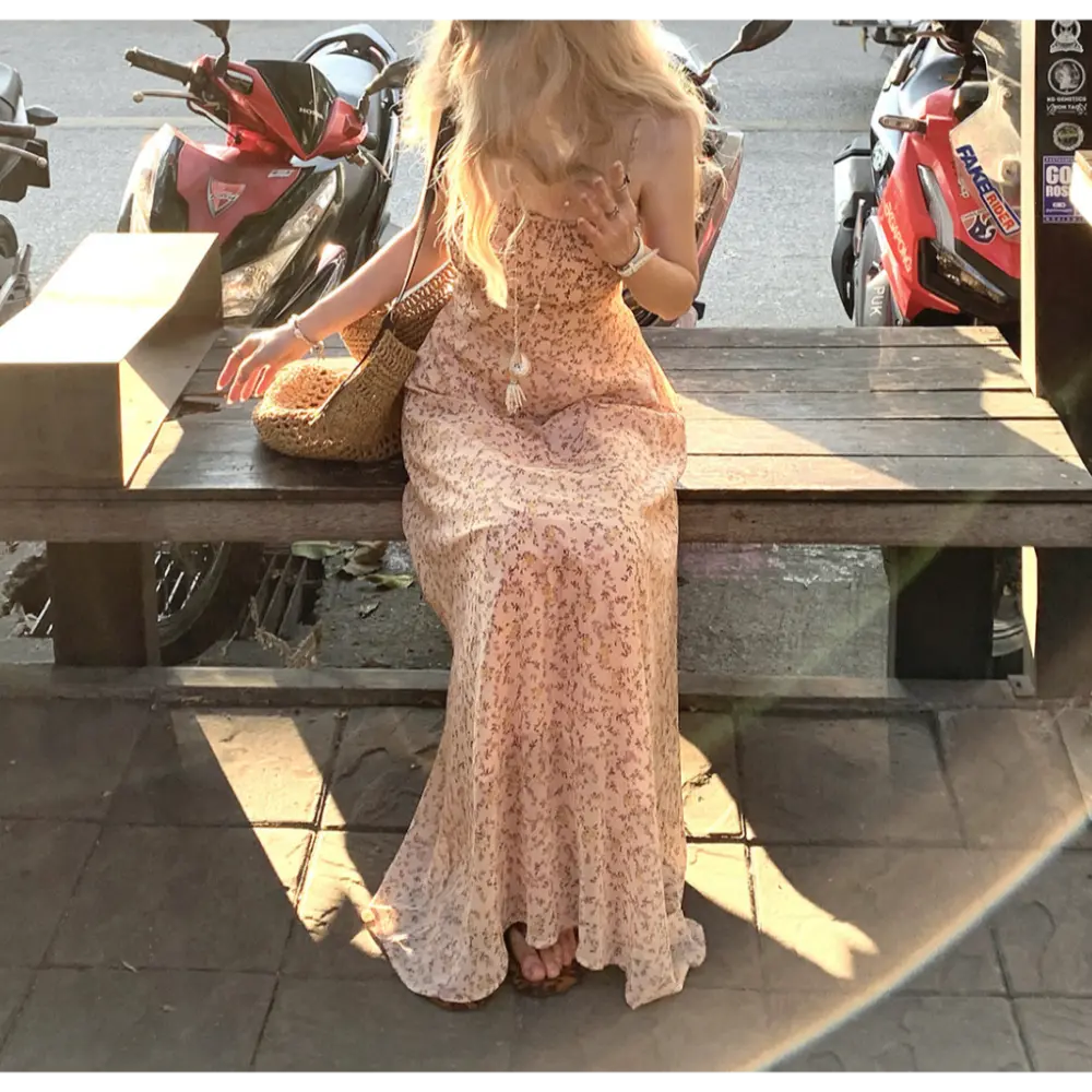 Sitting outdoor street lifestyle shot of light peach tiny floral maxi slip dress, model seated on dark wooden bench, rattan straw bag placed beside thigh, leopard print slide sandals visible