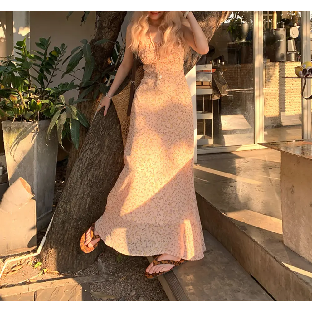 Golden hour outdoor courtyard lifestyle shot of light peach ditsy floral maxi slip dress, model leaning against tree trunk, wearing leopard print slide sandals, accessorized with woven straw shoulder bag
