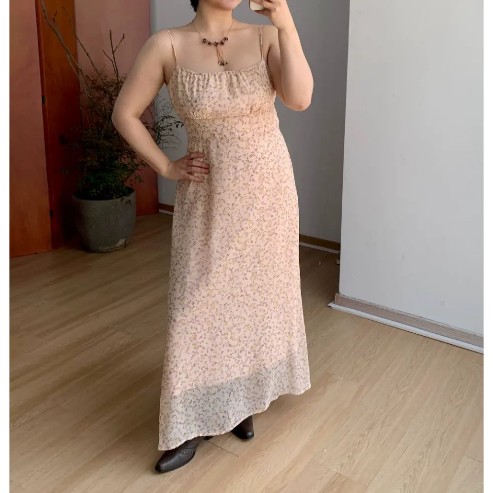 Straight front indoor mirror lifestyle photo of light apricot tiny floral maxi slip dress, gathered elastic bust, slim fitted waist, model resting one hand on hip with brown leather ankle boots
