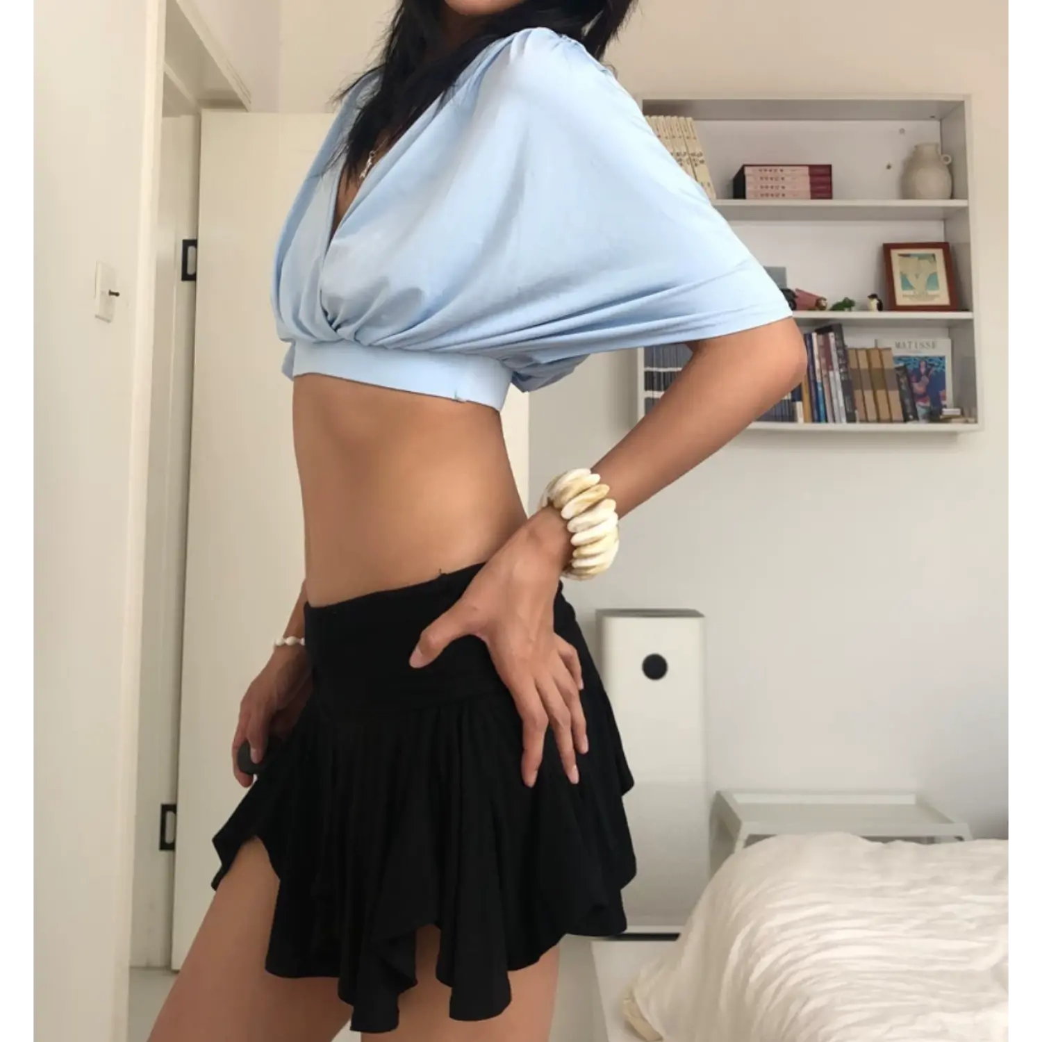 Three-quarter indoor bedroom lifestyle photo of surplice dolman sleeve crop top, model resting one hand on black ruched mini skirt to show waist shaping, loose batwing sleeves, sky blue matte jersey fabric, soft natural room lighting, plain white wall decor