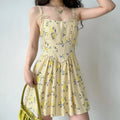 Woman wearing a pale yellow floral print spaghetti strap mini dress with tie shoulder straps, structured corset-style bodice, and flared skirt, styled with a green handbag.
