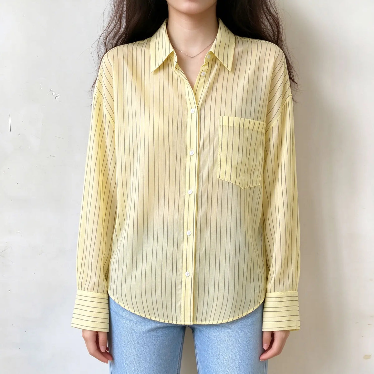 Model wearing pale yellow vertical stripe oversized button down shirt, collared neckline, chest pocket, long sleeve, loose fit, paired with light blue jeans against plain white background
