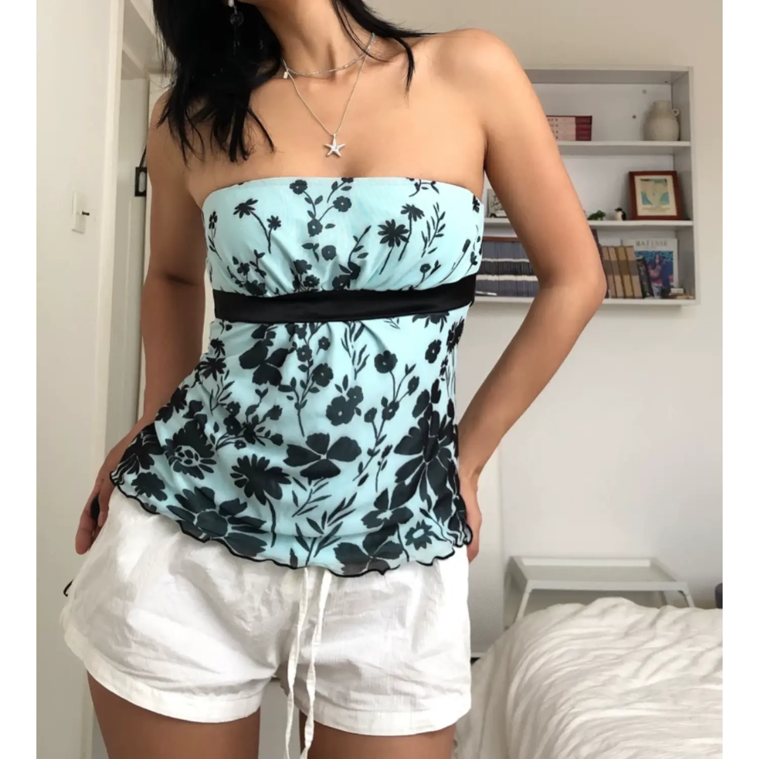Three-quarter indoor bedroom lifestyle shot of sheer mesh strapless tube top, soft ruffled wavy hem edge, wide black satin waist ribbon, model placing both hands on hips, styled with relaxed white drawstring shorts, bright clean white home decor background