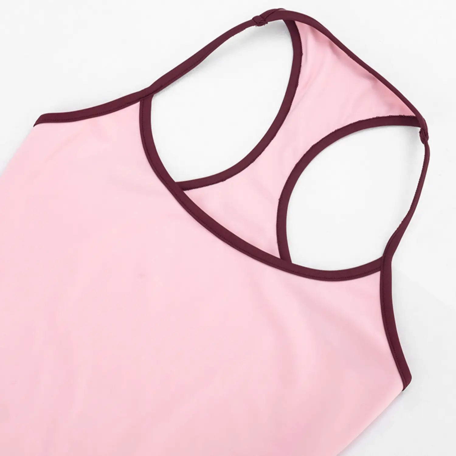 Partial angled close-up of halter tank rear racerback cutout, smooth matte pink stretch knit fabric, thin burgundy piping outlining neck and strap edges, clean tight seam construction