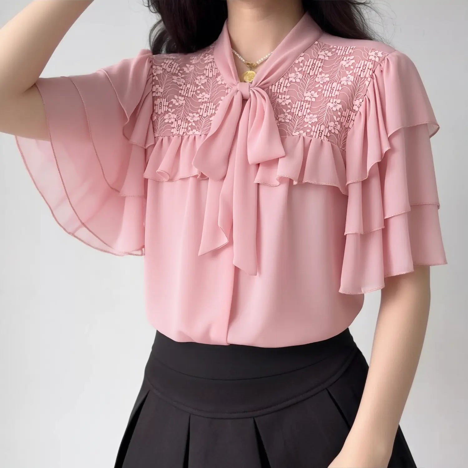 Lifestyle model outfit shot pastel pink chiffon blouse, tie bow neck, floral lace shoulder panel, tier flutter short sleeve