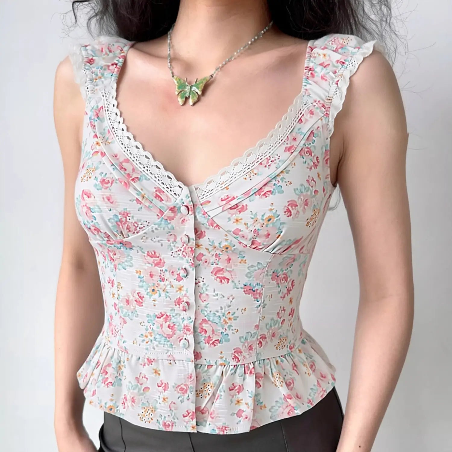 Model wearing cropped floral cami top, white base pastel pink blue yellow watercolor ditsy rose print, scalloped hollow lace trim deep V neck, ruffle elastic cap sleeves with lace edging, vertical self-fabric covered button placket, corset seamed fitted waist, flared peplum ruffle hem, slim vintage cottagecore silhouette