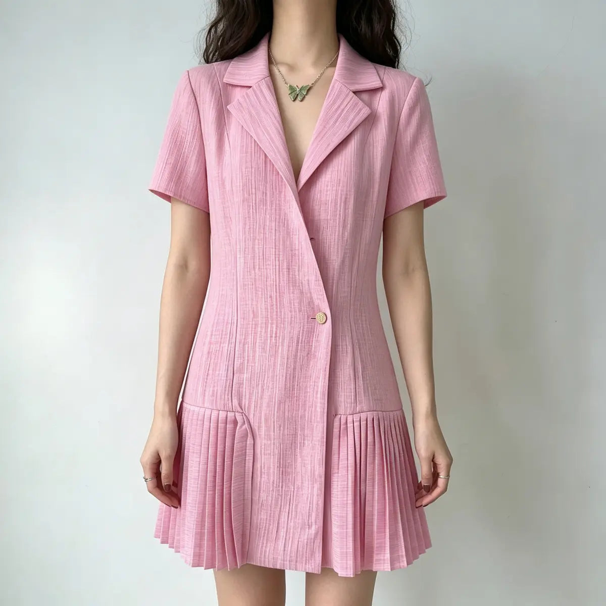 Model wearing a pink blazer-style mini dress with short sleeves, a single gold button, and pleated skirt hem, styled with a green butterfly necklace, front view.