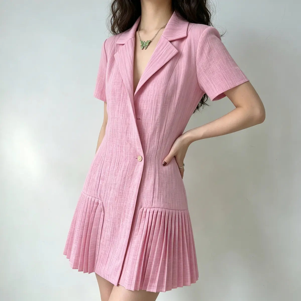 Model wearing a pink blazer-style mini dress with short sleeves, a single gold button, and pleated skirt hem, styled with a green butterfly necklace, side view.