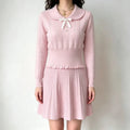 Model wearing pink cable‑knit two‑piece outfit set, scallop peter pan collar sweater with bow button detail and pleated mini skirt, front full view against plain wall