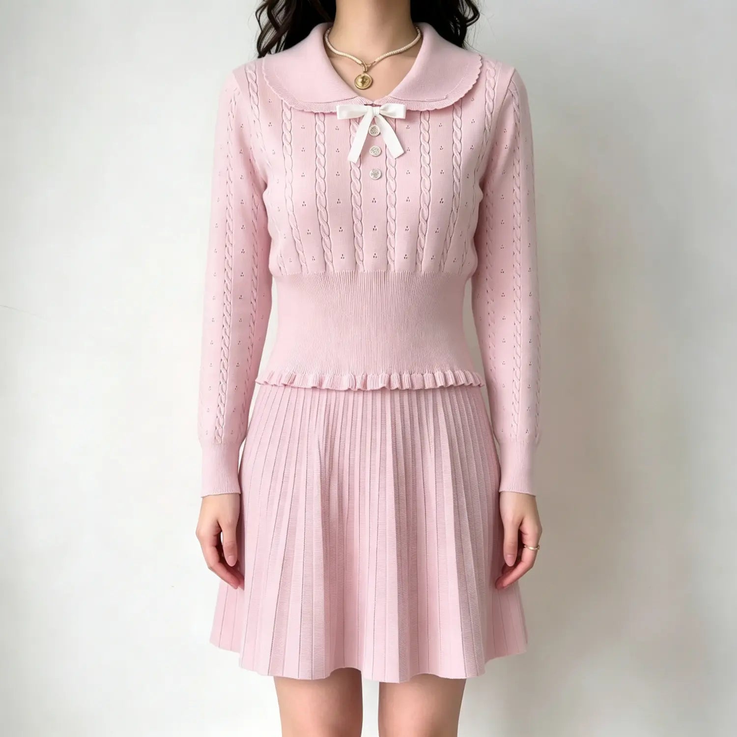 Model wearing pink cable‑knit two‑piece outfit set, scallop peter pan collar sweater with bow button detail and pleated mini skirt, front full view against plain wall