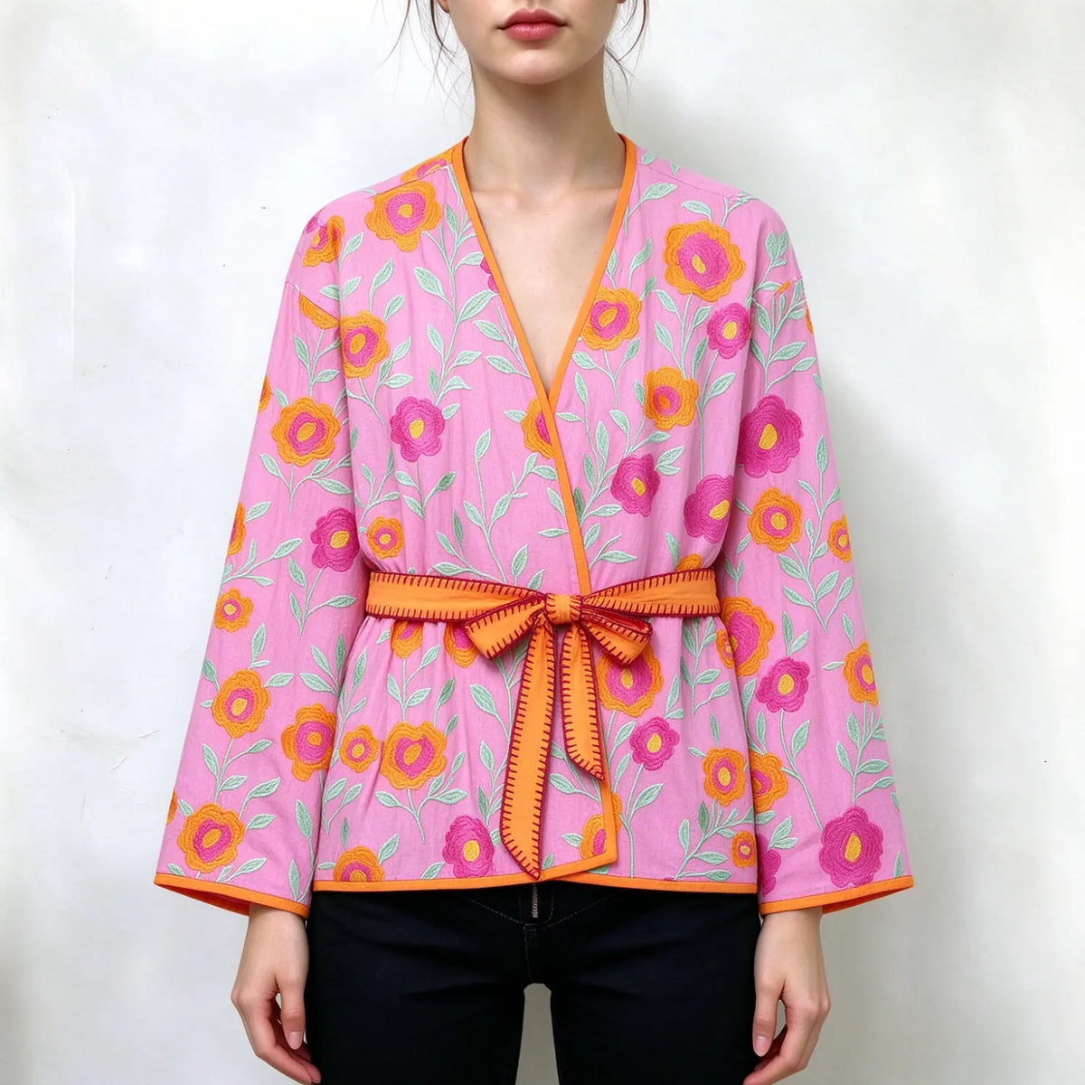 Model wearing pink wrap jacket covered with colorful floral embroidery, V neckline, orange trim, matching waist tie belt, flared long sleeves, loose fit, paired with black jeans, plain white background