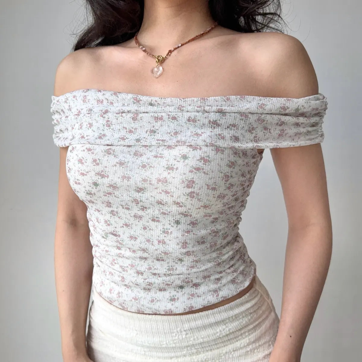 Front view of a woman wearing a pink floral off-shoulder top with a wide neckline, styled with a heart necklace and a white skirt.