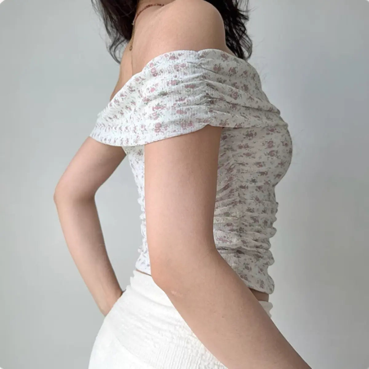  Side view of a woman wearing a pink floral off-shoulder top with gathered sleeves and side ruching, paired with a white skirt.