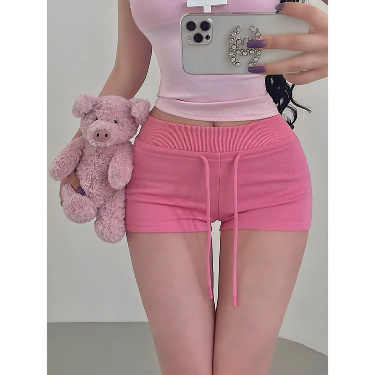 Close-up of a woman wearing pink high-waisted drawstring shorts with a matching top, holding a pink teddy bear.