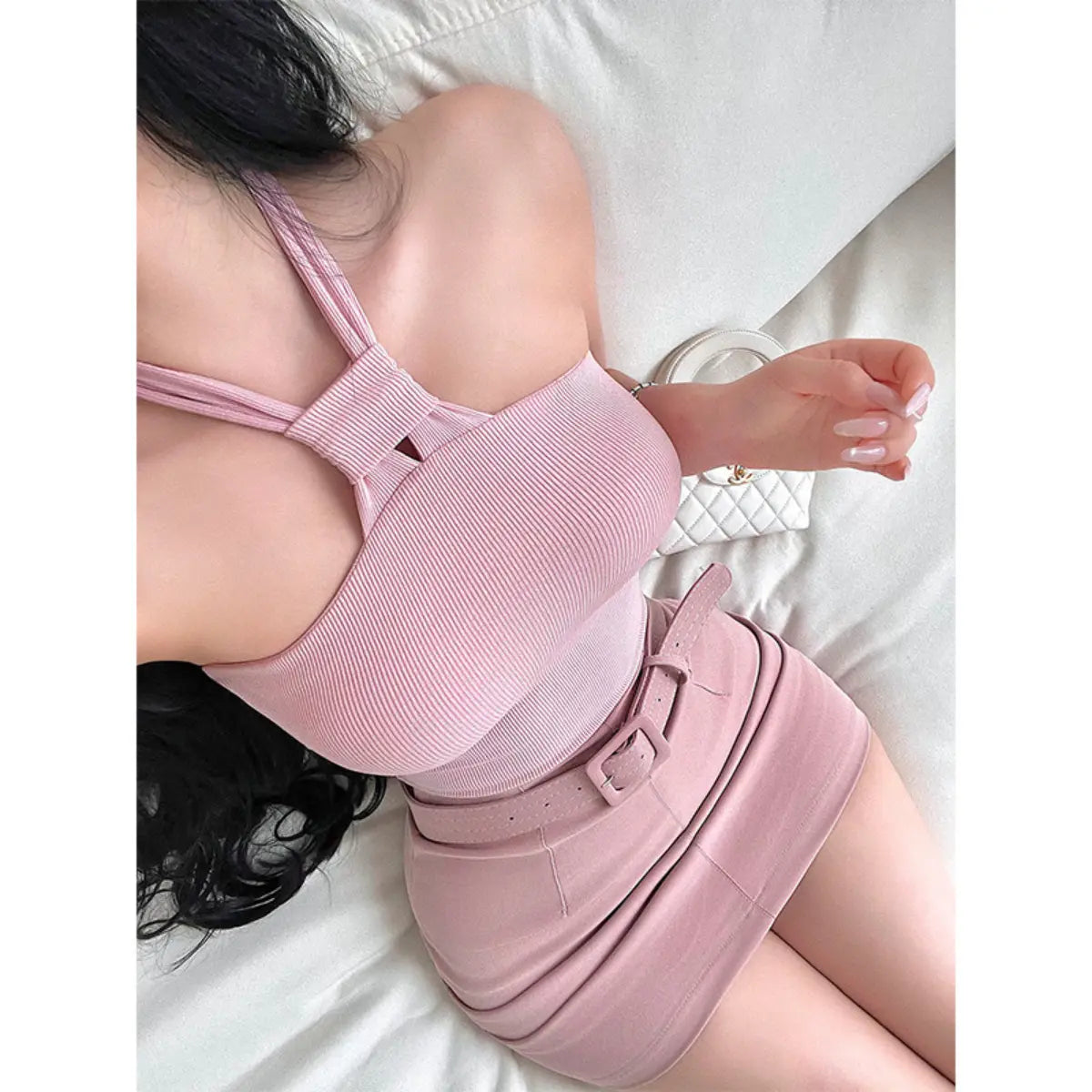 A woman wearing a pink ribbed keyhole halter top, styled with a matching belted mini skirt, taking a bed selfie.