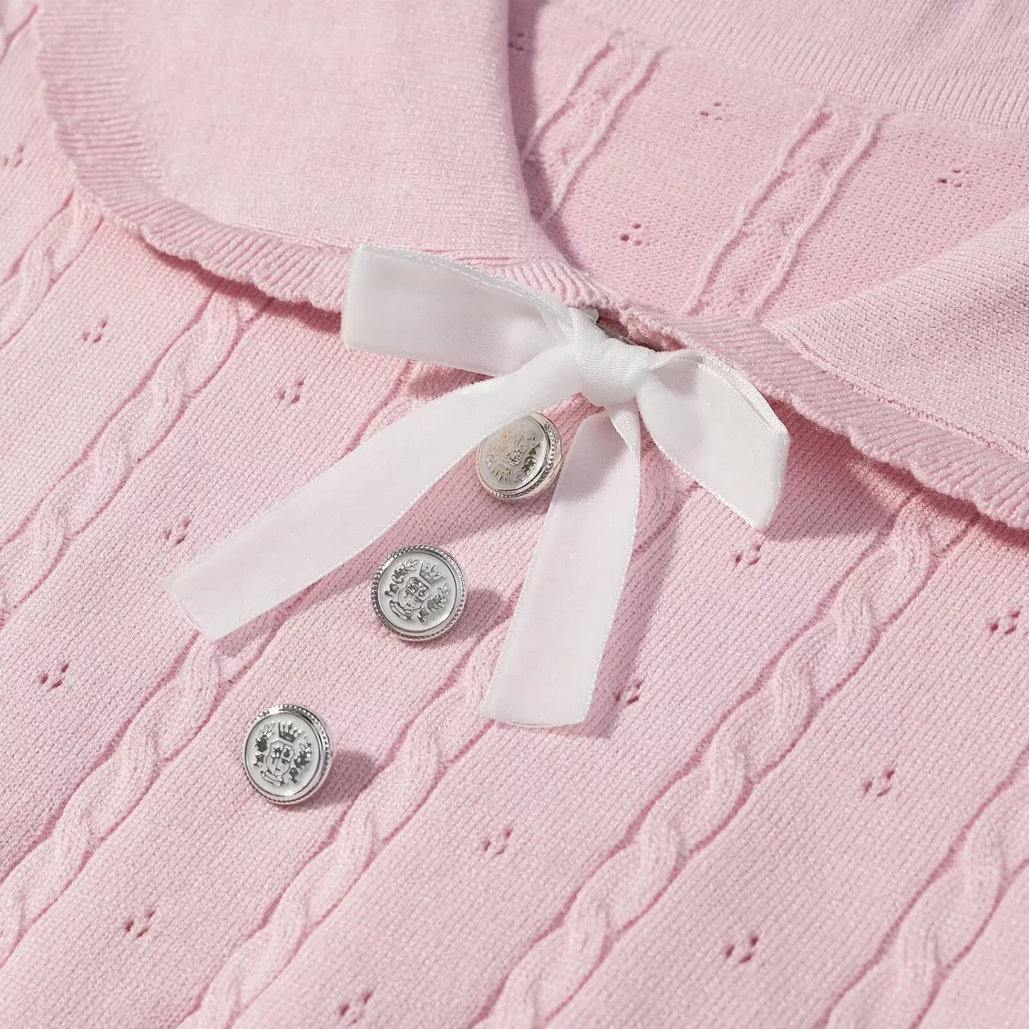 Macro close‑up of white satin bow and engraved silver decorative buttons on pink cable knit sweater