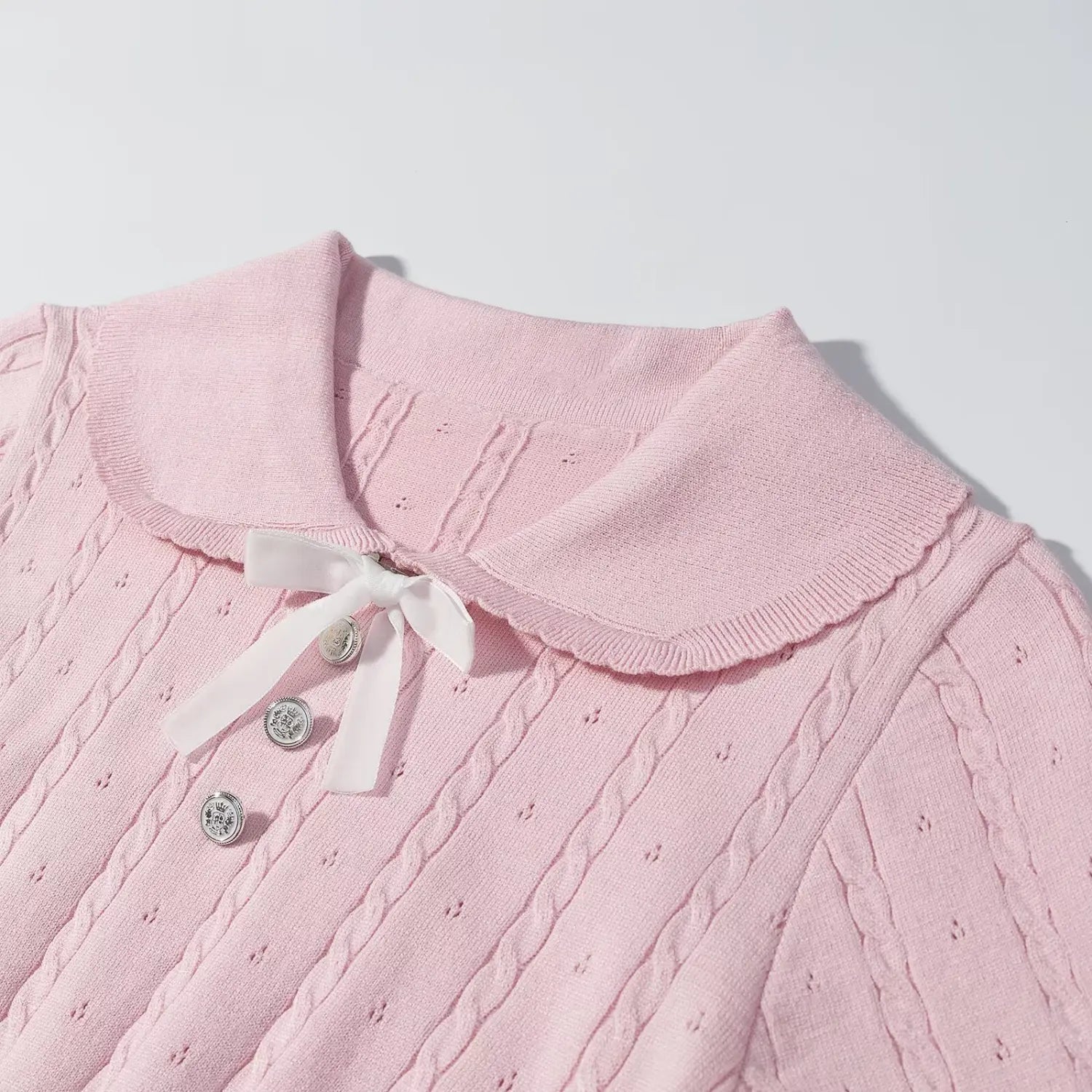 Close‑up detail of pink knit sweater scalloped peter pan collar, white satin bow tie and engraved silver buttons, cable knit fabric texture