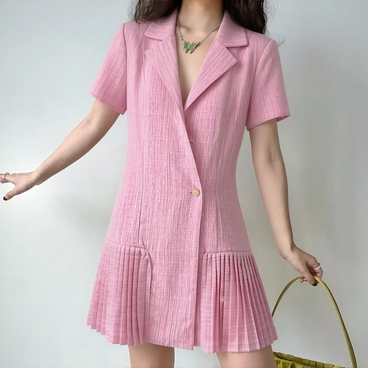 Model wearing a pink linen blazer dress with a single gold button, styled with a green bag and butterfly necklace.