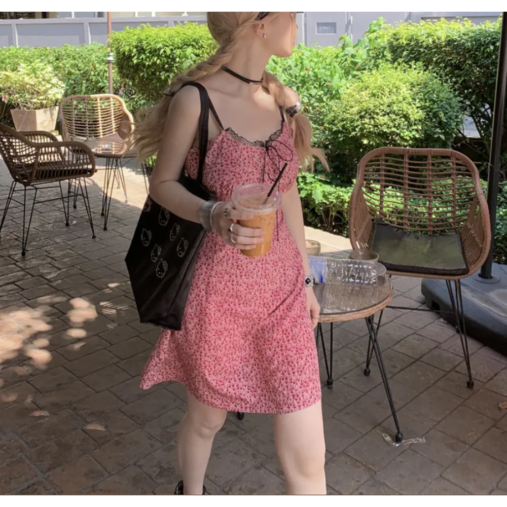 Side profile outdoor cafe lifestyle shot of pink ditsy floral cami mini dress, model holding iced coffee cup, accessorized with black canvas tote bag and silver wrist bangles