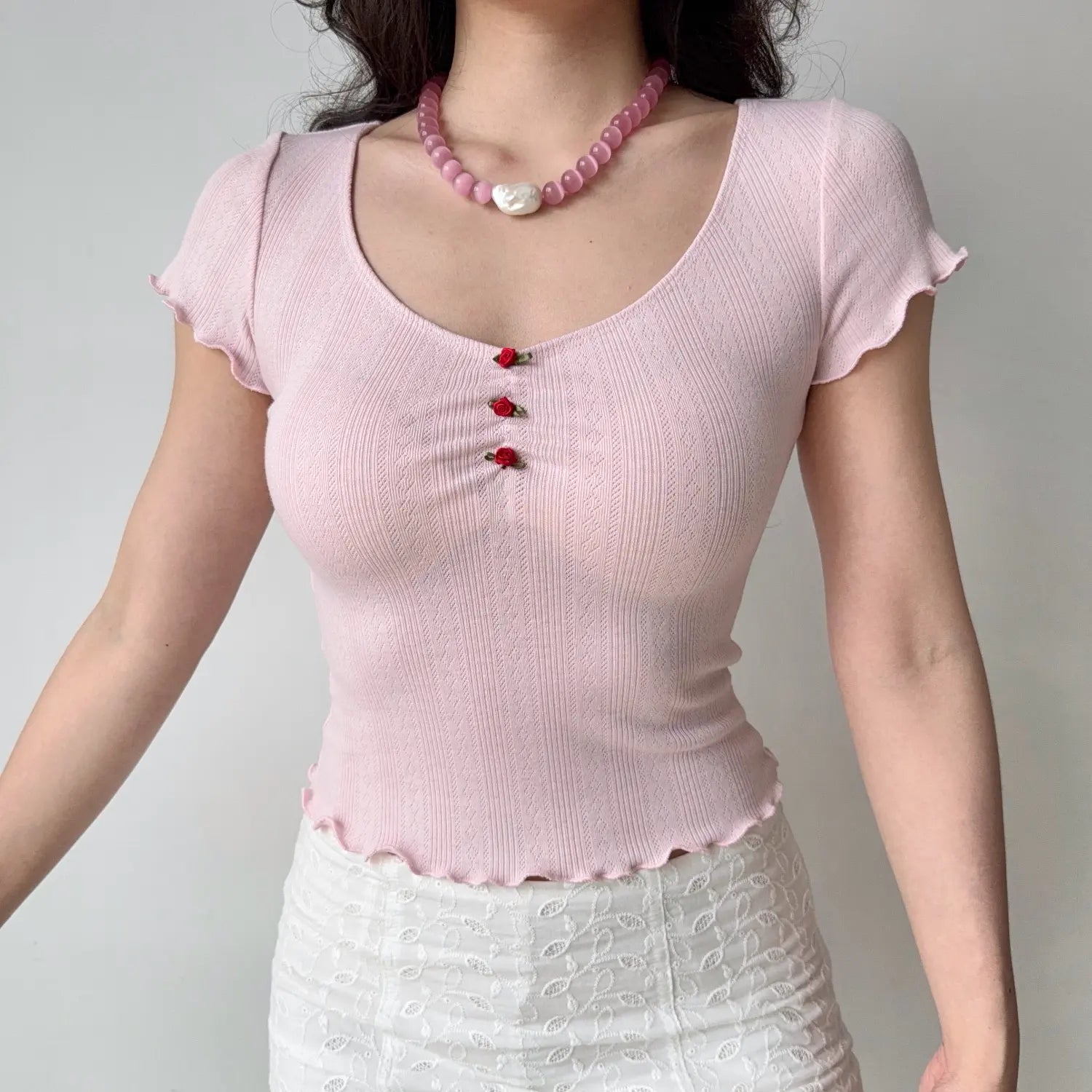 Front view of model wearing the pink ribbed knit top with rose bud buttons, paired with a white lace mini skirt