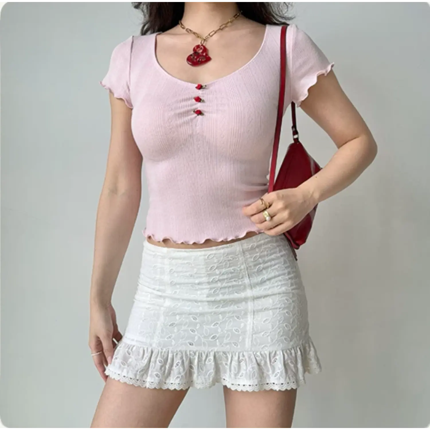  Lifestyle shot of model in pink rose-detail knit top with red shoulder bag, styled with a white lace skirt