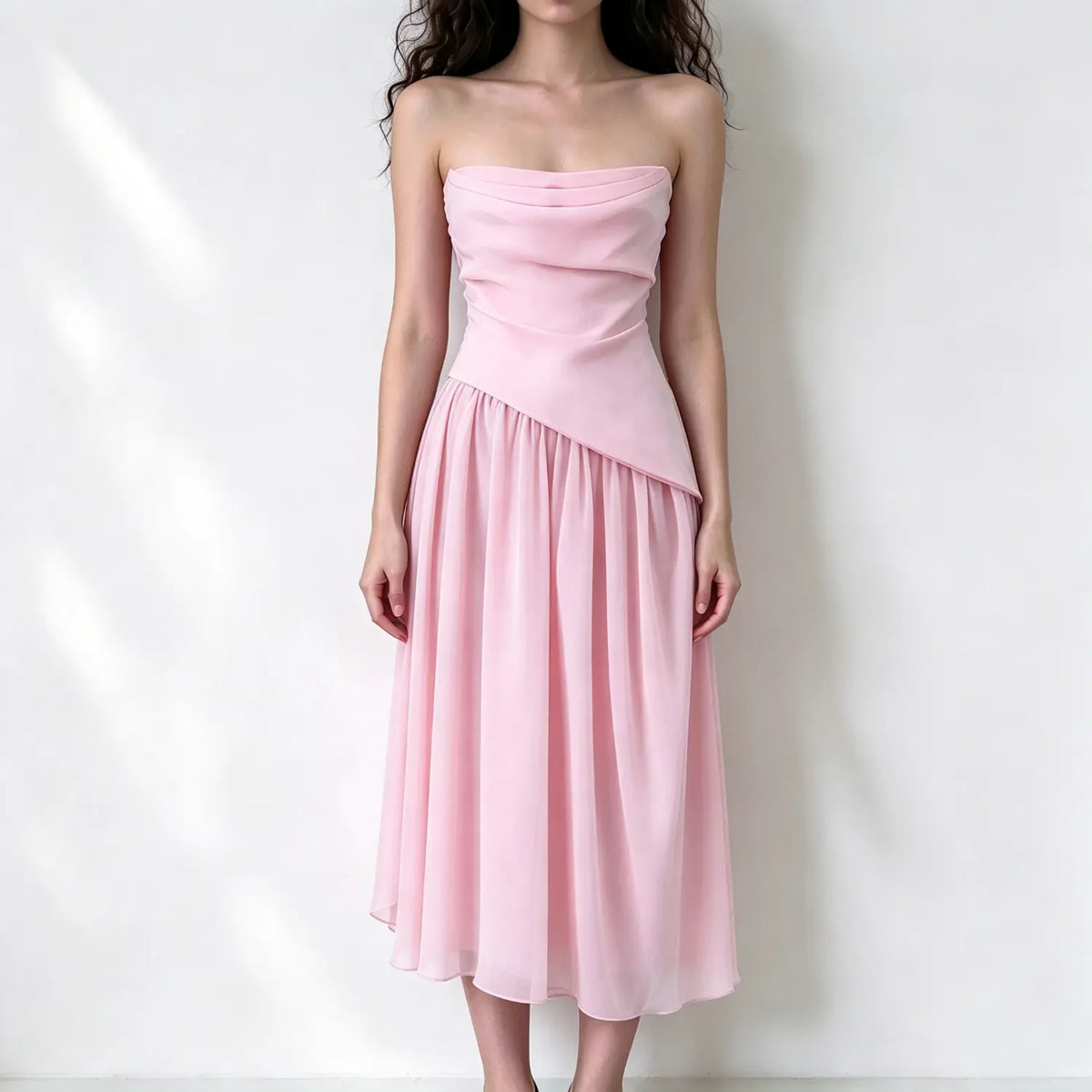 Model wearing soft pink strapless midi dress, draped ruched bodice with asymmetrical hem, flowy chiffon pleated skirt, plain white background