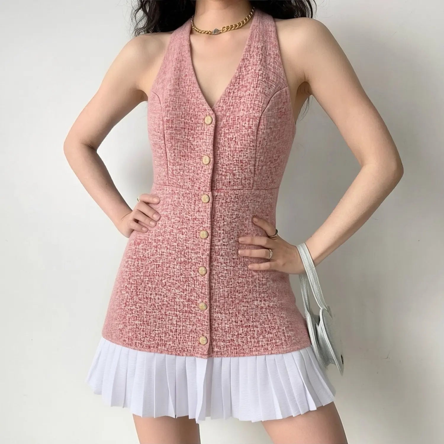 Model daily shot of heather pink tweed mini dress, sleeveless halter deep V neck, vertical row gold embossed buttons, fitted waist, contrast white box pleat hem, styled with thick chain gold necklace & light gray mini handbag, sweet elegant date look