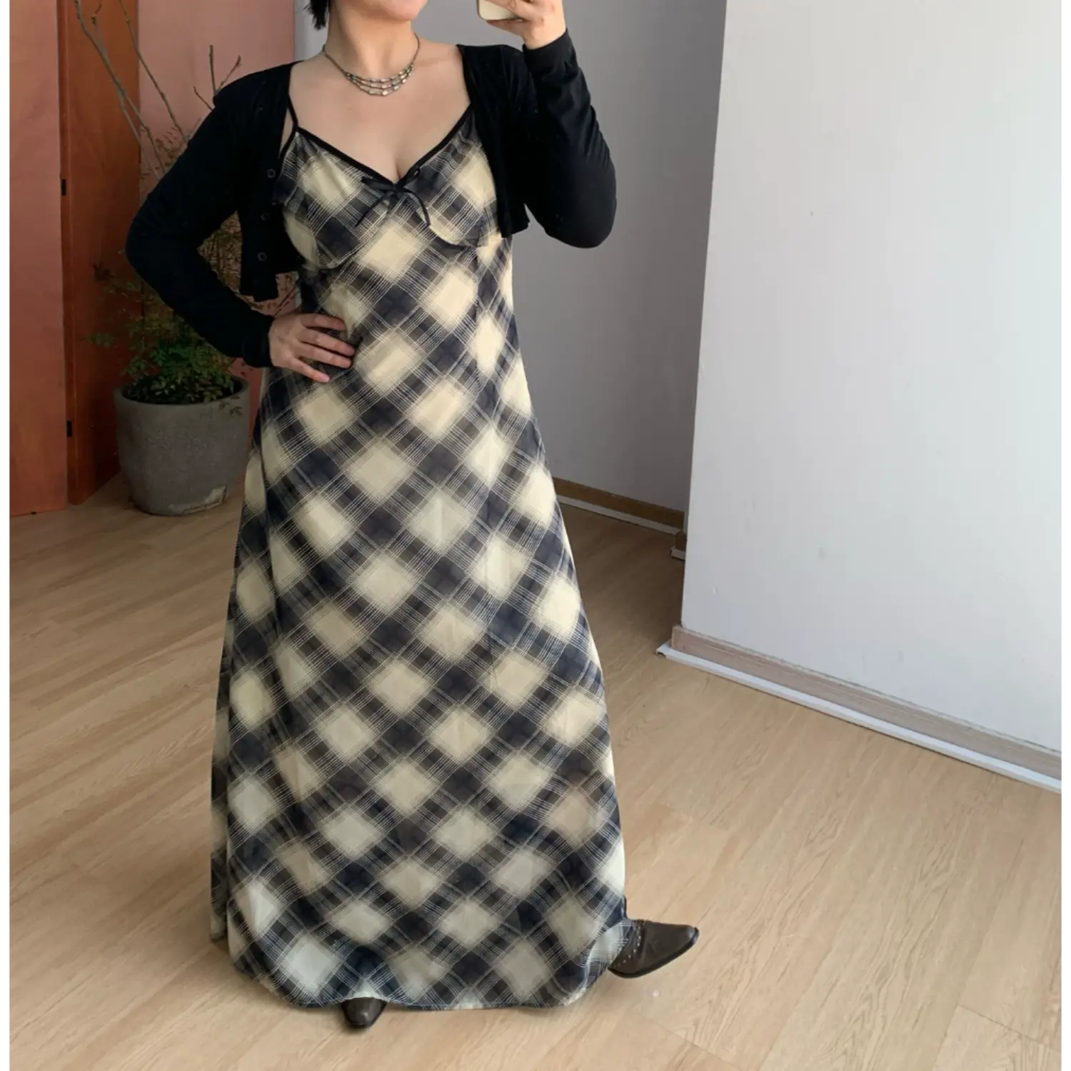 Indoor mirror styling shot of diagonal plaid slip maxi dress matched with cropped long sleeve black knit cardigan, model resting one hand on hip to show waist shaping, contrast black neck piping, tiny front bow detail, brown leather low heel shoes, simple potted plant decor