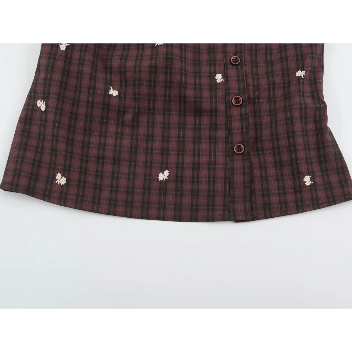 Close-up of curved hem of plaid vest with three round reddish brown buttons with black buttonholes and floral embroidery