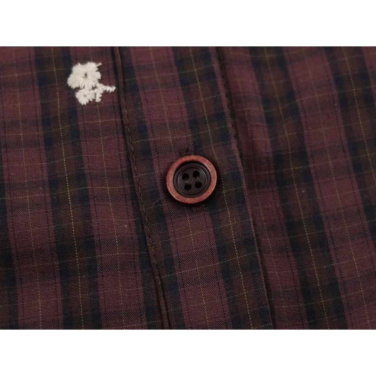 Close-up of front placket with four-hole reddish brown button and white floral embroidery on plaid fabric