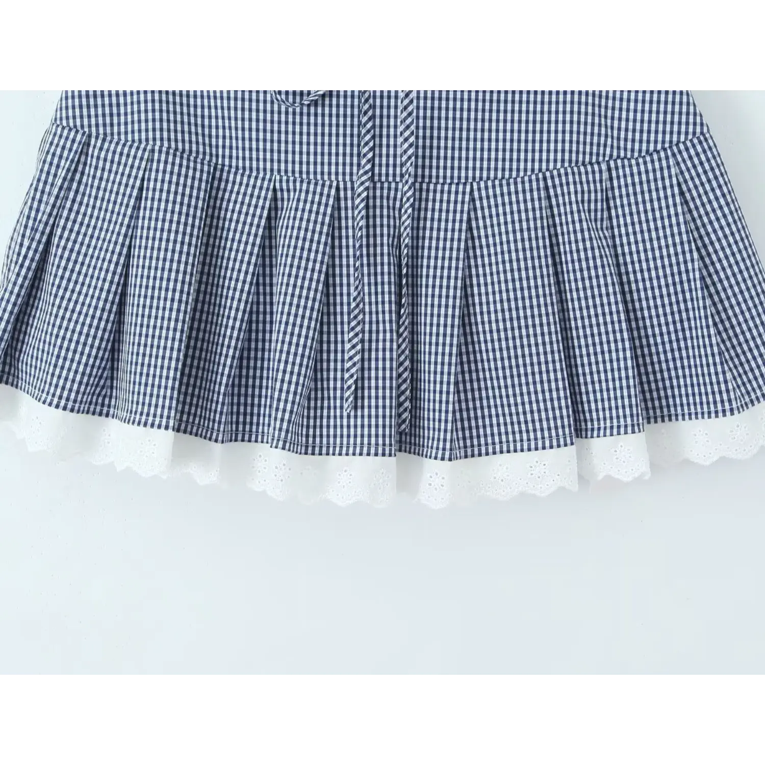Navy gingham pleated mini skirt hem flat detail shot, uniform three-dimensional box pleats full circle white eyelet wavy lace hem waist matching check drawstring tie high waist A-line skirt silhouette display
