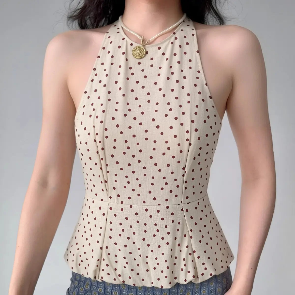 Model wearing a beige halter peplum top with red polka dots, showcasing the high neck, fitted bodice, and flared peplum silhouette.