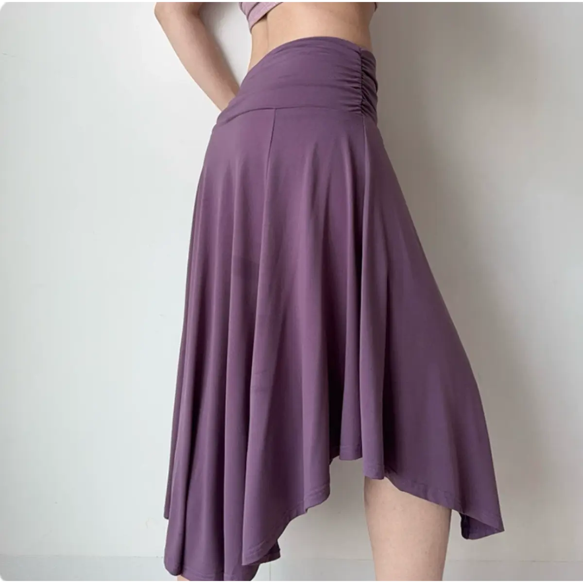 Side view of a purple asymmetrical midi skirt with a ruched waistband and flowing, uneven hem, worn with a cropped top.