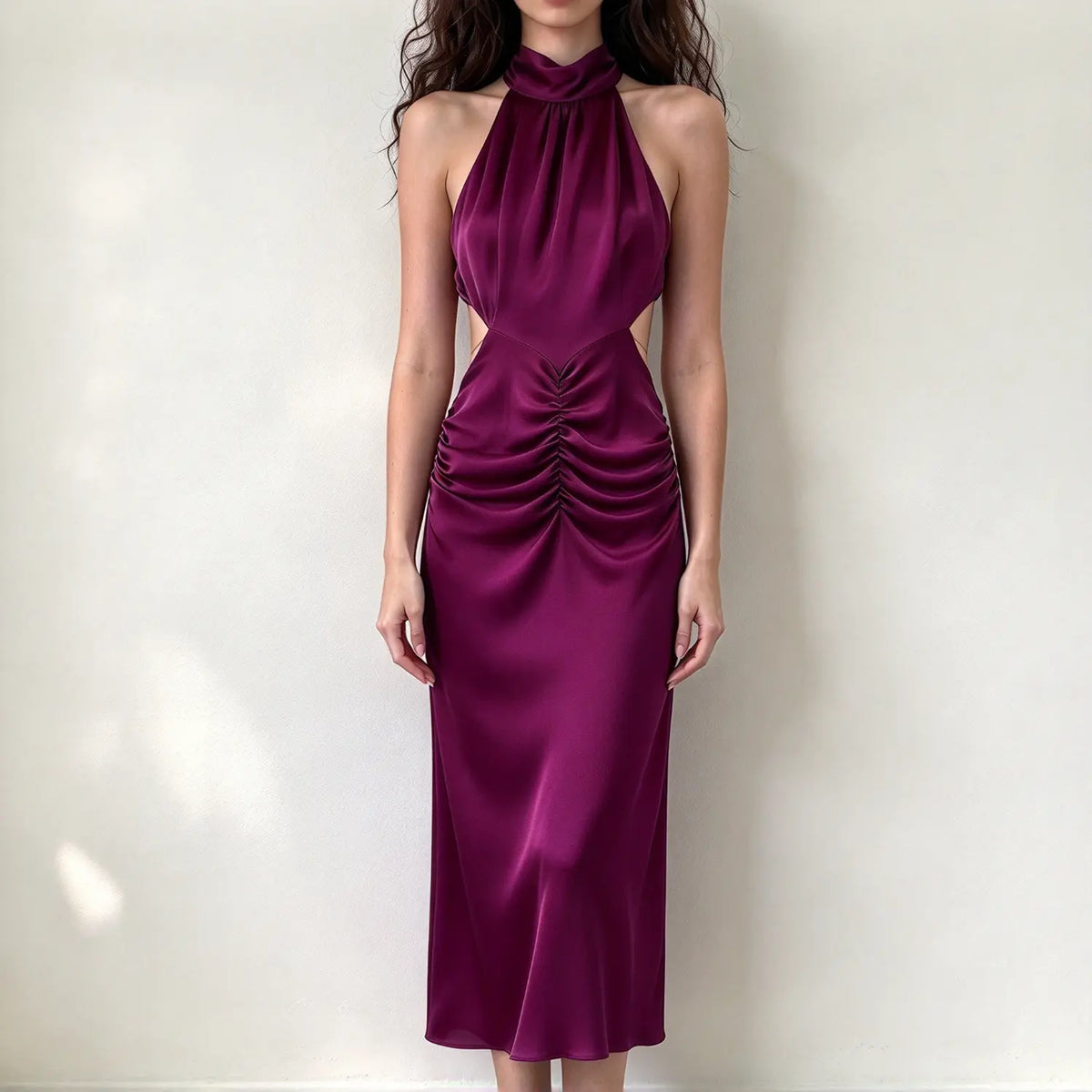Model wearing deep purple satin halter maxi dress, high neck, side waist cutouts, front ruched detail, sleek flowing skirt, white backdrop