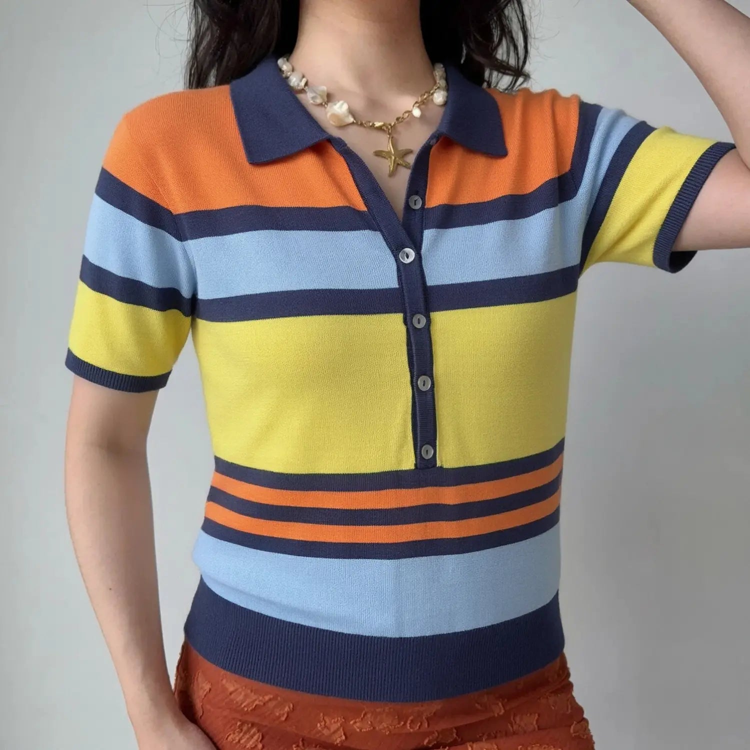 Lifestyle model wearing striped knit polo top, navy lapel collar, half shell button placket, slim fit silhouette, matching casual bottom outfit, retro color block rainbow stripe design