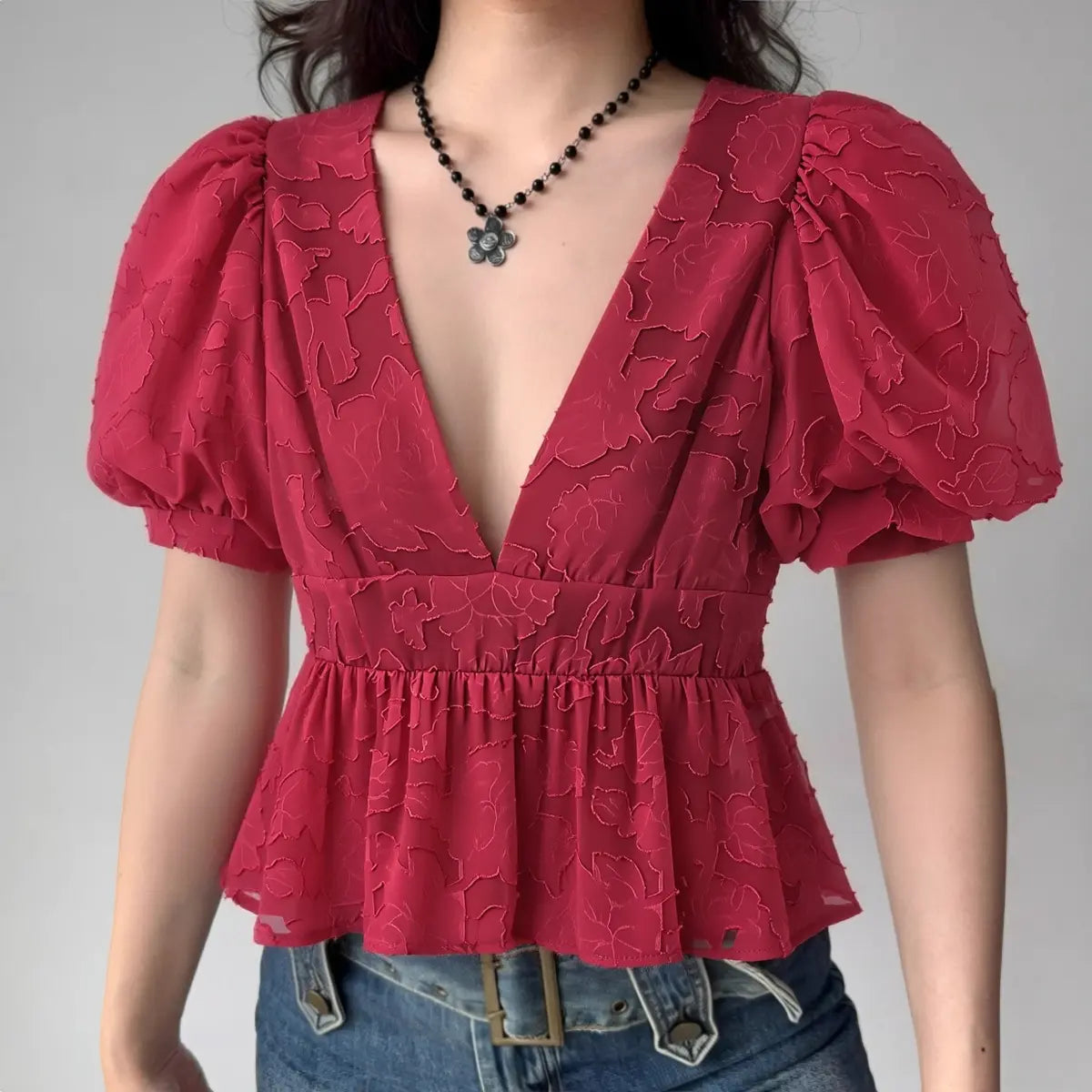 Woman styled in a red floral appliqué peplum top, featuring a deep V-neck, puff sleeves, and gathered waist, paired with blue jeans.