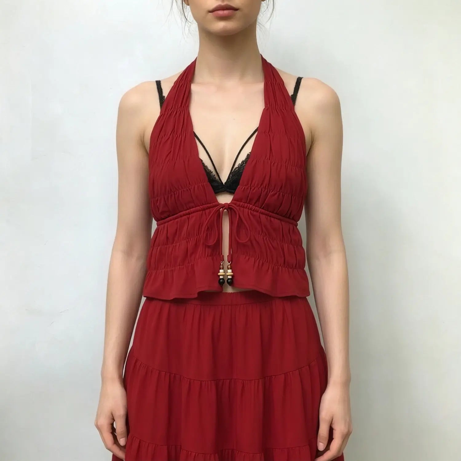 Female model wearing red two‑piece outfit, deep v halter ruched crop top and tiered ruffle maxi skirt, plain light studio background