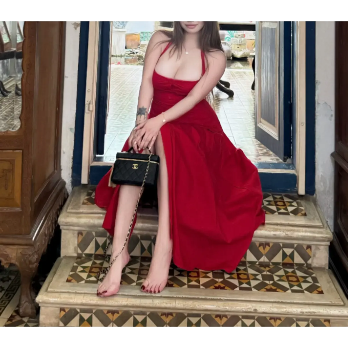 Woman in a bright red halter maxi dress sitting on a tiled staircase, holding a black handbag, with bare feet and red nail polish.