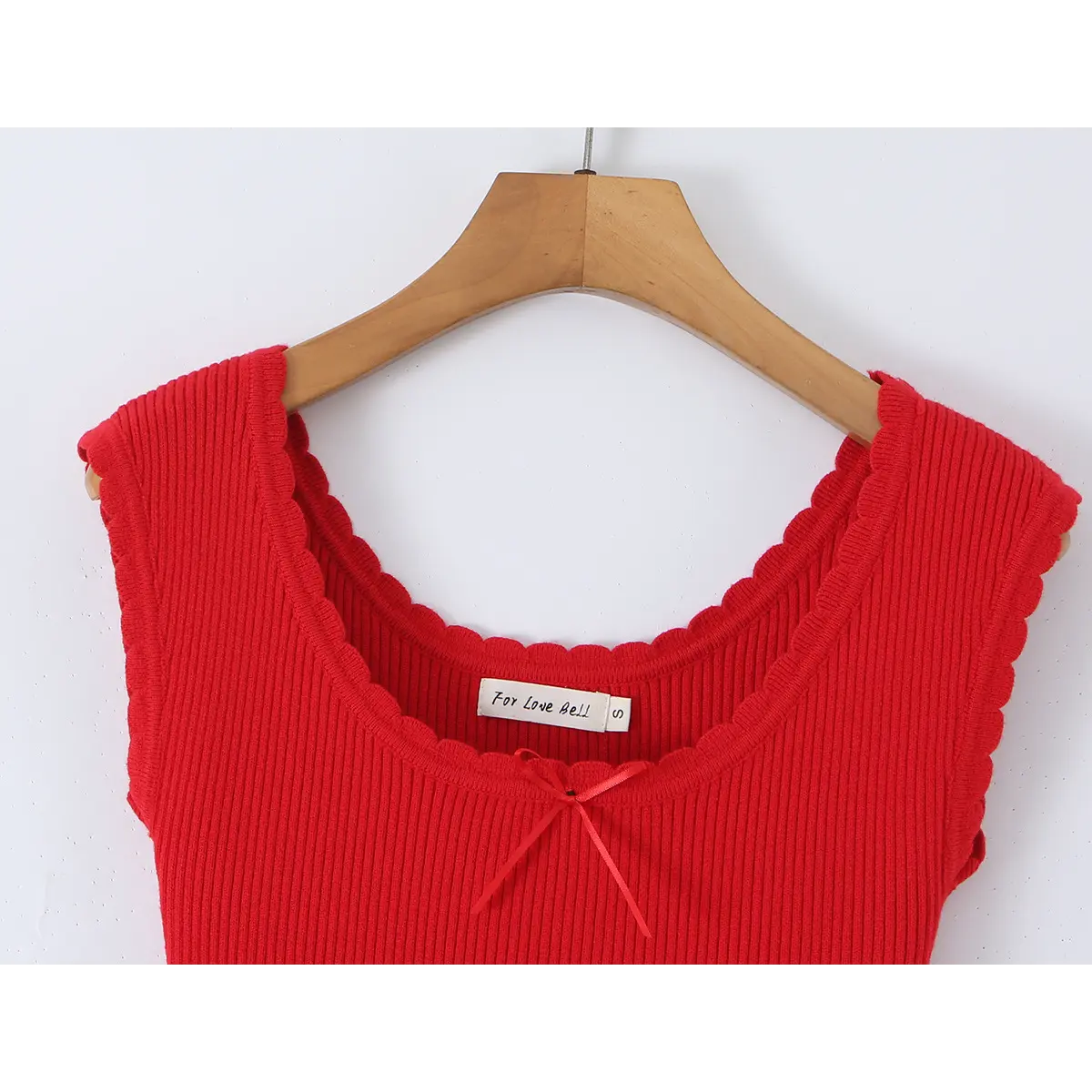 Close-up of the neckline on a red knit dress hung on a wooden hanger, showing the scalloped edge, center ribbon bow, and brand label.