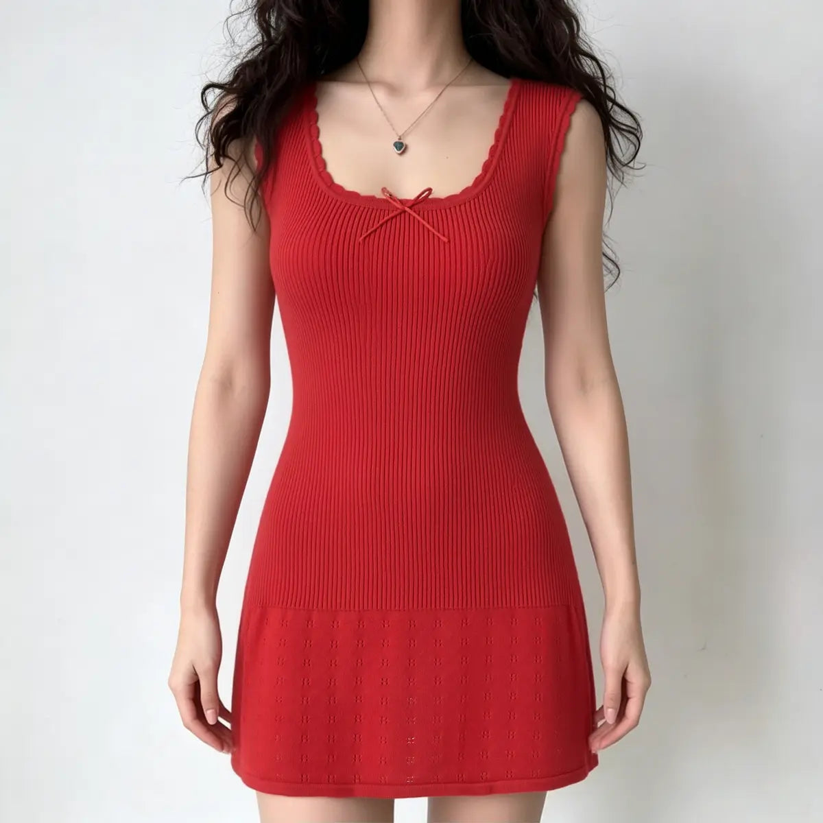 A woman wearing a red ribbed knit mini dress with scalloped neckline, cap sleeves, center ribbon bow, fitted bodice, and flared eyelet-knit skirt, styled with a heart pendant necklace.